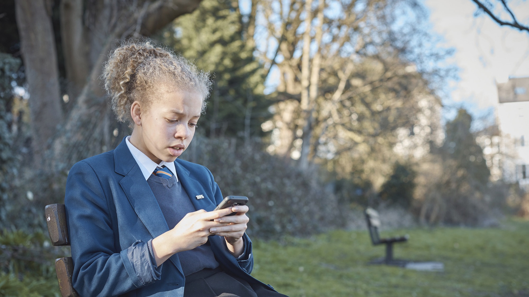A teenage girl in school uniform on her mobile while sitting on a park bench.