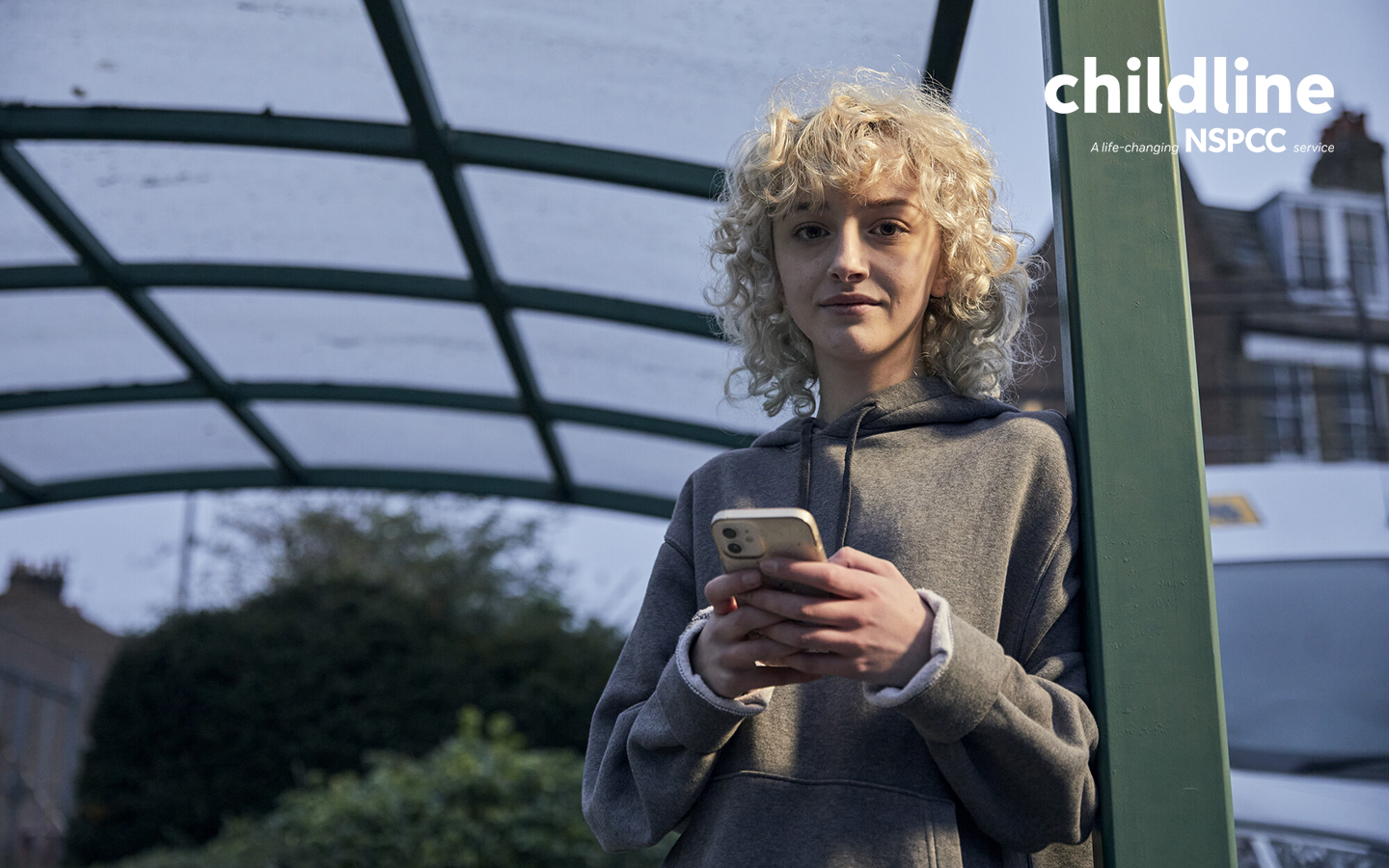 A teenage girl using her phone outside a bus stop while smiling into the camera. A white NSPCC and Childline logo lockup is layered into the top right corner of the image which says 'Childline: a life-saving NSPCC service'.