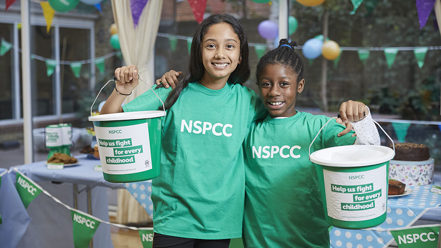 Two teenage girls in NSPCC t-shirts smiling while holding fundraising buckets.